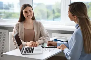 Two women sitting at a table, one holding a pen and the other holding a laptop, looking at each other and smiling in an office