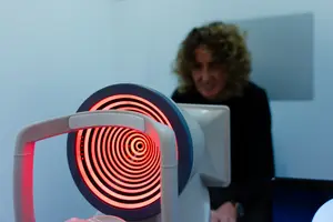 An older woman with curly hair is standing behind a machine with a red spiral light and looking at a monitor.
