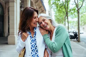 Two women standing on a sidewalk, one with her hand on the other's shoulder, both smiling