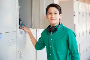 A smiling boy with headphones on his neck is standing in front of a locker, his hand on the door handle.