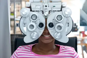A woman is sitting on a chair in front of an eye examination machine, smiling and looking at the camera.