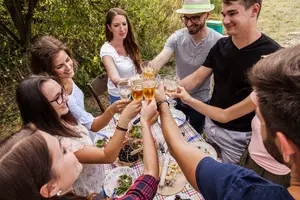 A group of people sitting around a table outdoors, some holding up glasses of beer and toasting to each other, with food and drinks on the table.