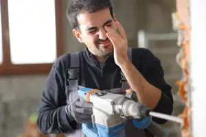 A construction worker using a drill machine while covering his eyes with his hand.