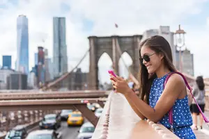 A smiling woman on a bridge looks at her pink phone with cars below her.