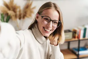 A woman in glasses smiles at the camera while standing indoors next to a bookshelf.