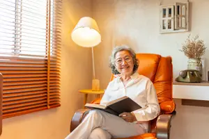 A smiling elderly woman wearing glasses sits in a comfortable chair reading a book in a cozy room