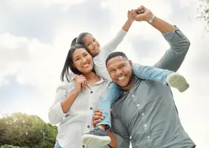 A family in a park with a smiling girl on the father's shoulders, the mother holding her waist, and a cloudy sky in the background.