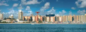 A panoramic view of a cityscape with tall buildings and a red tower, under a blue sky with clouds, with a body of water in the foreground.