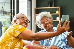 Two elderly women sitting on a couch, one of them is taking a selfie using a smartphone, and they are both smiling and looking at the camera