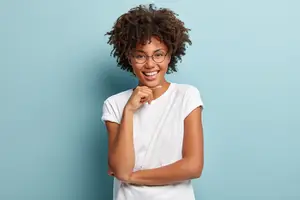 A smiling woman wearing a white t-shirt and glasses with curly hair, posing for a photo against a blue background.