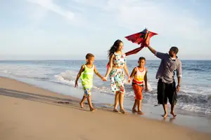 A family of four, including two boys and two adults, are walking on the beach at sunset, holding hands and a kite.