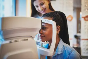 A young woman is getting her eyes checked at an optometrist office with another woman smiling in the background.