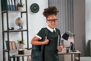 Young girl in school uniform with backpack and books posing for a photo in a classroom