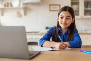 A young girl is sitting at a wooden table with a laptop, notebook, and pen on it, and she seems to be doing her homework.