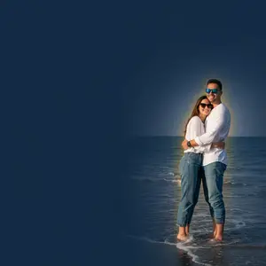 A couple standing in the water, smiling at the camera, on a beach with a blue sky and ocean in the background