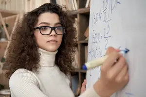 A young woman wearing glasses and a white turtleneck shirt is writing on a whiteboard with a marker.