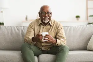A smiling senior African American man sitting on a couch, holding a white coffee cup, wearing eyeglasses and a beige shirt, with a white wall and potted plant in the background.