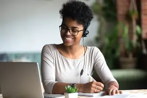 A smiling woman wearing a headset and glasses is sitting at a table with a laptop and a notebook in front of her, probably working on a project.