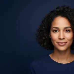 Portrait of a smiling woman with curly hair and brown eyes, wearing a blue shirt, standing against a dark blue background.