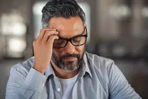 A man with a beard and glasses is sitting in an indoor area and holding his forehead with his right hand, probably feeling headache or tired.