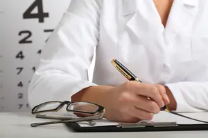 A doctor sits at a desk and writes in a book while wearing a white lab coat.