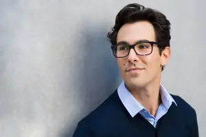 A smiling man wearing glasses and a blue sweater is leaning against a gray wall.