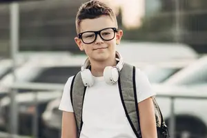 A boy wearing glasses, a white shirt, and a backpack with headphones is standing in front of a parking lot.