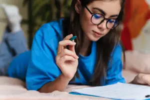 A young girl with glasses is reading a book on a bed and holding a pen.