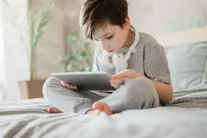 A boy with headphones sitting on a bed holding a tablet