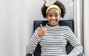 A smiling girl sitting in a chair wearing a yellow headband and a striped shirt