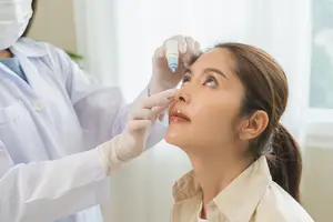 A doctor in a lab coat is administering eye drops to a woman in a hospital room.