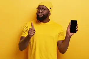 A man wearing a yellow shirt and hat is holding a smartphone and giving a thumbs-up gesture