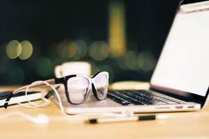 A laptop with a pair of sunglasses and headphones on a wooden table with a cup and a pen