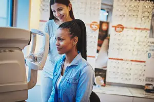 A woman is using an eye examination machine while another woman is smiling behind her, probably in an optical shop.