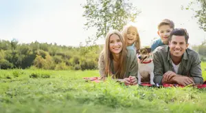 A family is lying on the grass in a park with a dog and smiling at the camera
