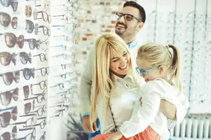 A family is inside an optical shop, the woman is holding a child and the man is smiling at the camera.