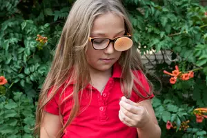 A girl in a red polo shirt with a pink collar is wearing glasses with pink lenses and looking at something in her hand, with plants and flowers in the background.