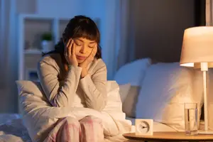 A girl sitting on a bed with pillows and her hand on her forehead while looking down