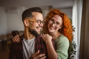 A man and a woman are smiling and posing for a photo in their home. The man has a beard and is wearing glasses. The woman has curly hair and is wearing a green sweater. Behind them is a white curtain and a plant. On the left side, there is a wooden table and chairs. On the right side, there is a window with a white curtain.