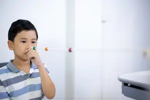 A boy with black hair blows through a long tube with colored balls on the ends, possibly in a medical setting