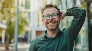 A man wearing glasses and a green shirt is standing in front of a building with his hand on his head and smiling.