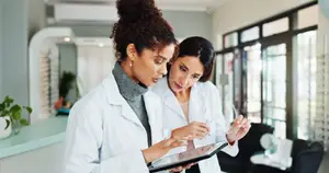 Two women in white lab coats are looking at a tablet in a modern medical office.