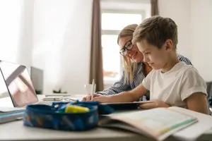 A woman and a boy are sitting at a desk and looking at a laptop. The boy is holding a pen and seems to be writing. The woman is smiling and looking at the boy. On the desk are a laptop, a book, a pen, and a bag. Behind them is a window with a curtain.