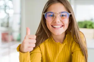 A young girl wearing glasses and a yellow sweater is smiling and giving a thumbs-up gesture in a room with a blurry view of a glass wall and plants outside.