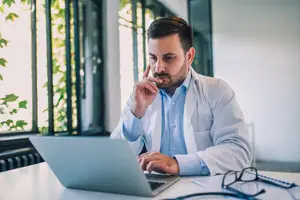 A man in a lab coat is using a laptop to check insurance benefits.