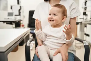 A baby is sitting on a chair and smiling while a woman is holding her and looking at her