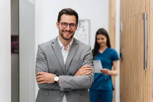 A man with glasses and a watch is smiling and standing in a hallway with a woman in blue scrubs standing behind him