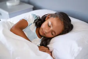 A young girl with long black hair sleeps peacefully in a bedroom, wearing orth-k contact lenses