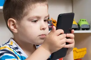 A young boy is looking at his phone while standing in front of a shelf with toy cars.
