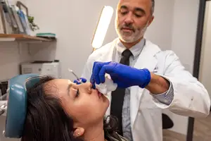 A doctor in a white coat and blue gloves is administering a nasal spray to a woman in a dental chair.